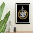 Framed artwork with Arabic calligraphy on a black background, placed on a wooden surface with decorative vases and a plant.