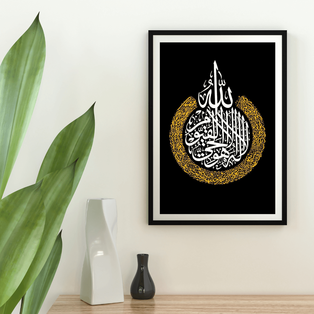 Framed artwork with Arabic calligraphy on a black background, placed on a wooden surface with decorative vases and a plant.