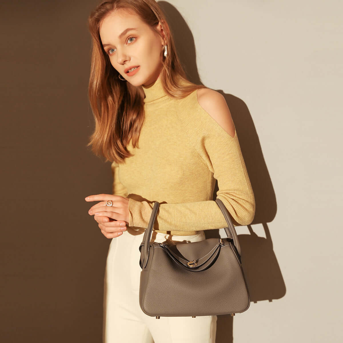 Woman in a yellow sweater holding a Monte Convertible Shoulder Bag, showcasing its elegant design and gold-plated hardware.