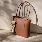 Brown leather tote bag with a teddy bear charm on a textured surface.