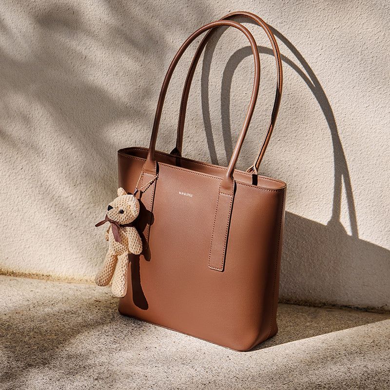 Brown leather tote bag with a teddy bear charm on a textured surface.