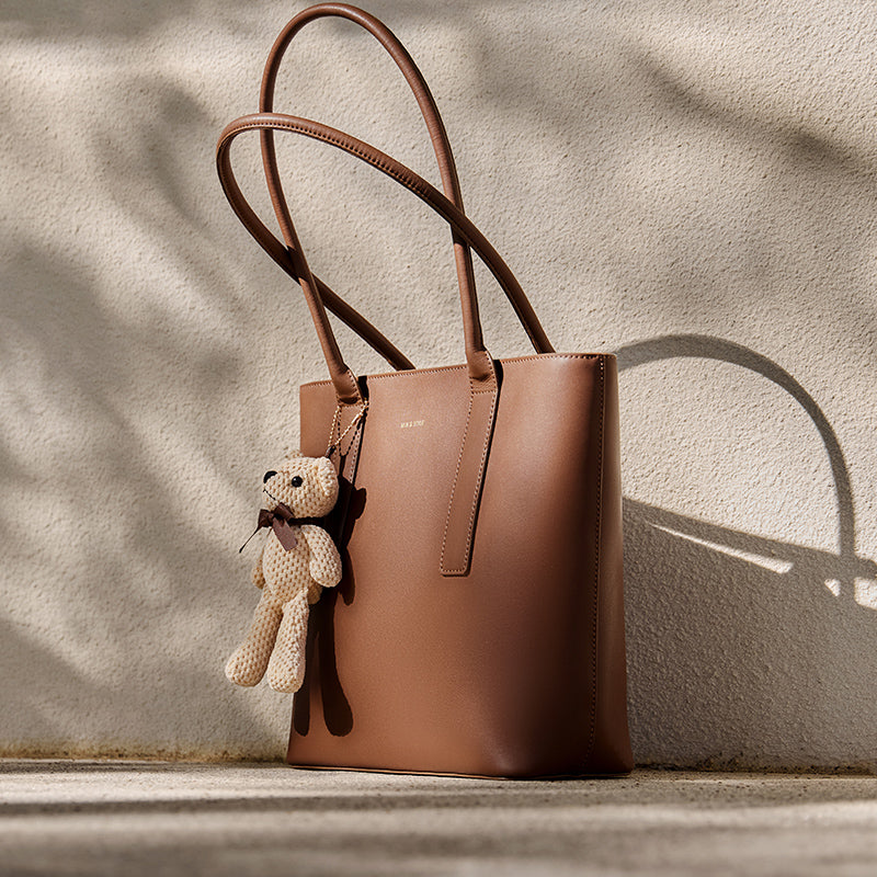 Brown leather handbag with a teddy bear charm on a neutral background