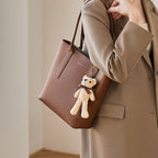 Brown handbag with a teddy bear charm held by a person wearing a beige coat.